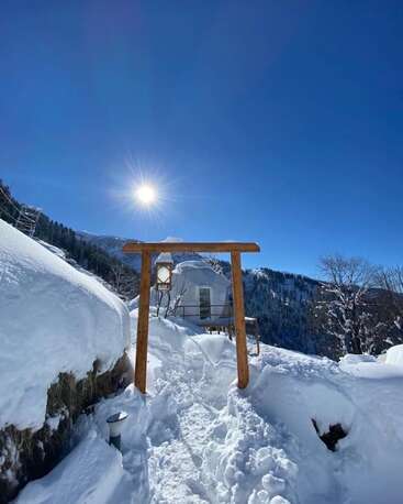 Un ciel bleu limpide illumine un paysage enneigé, où un portail en bois encadre un sentier menant à une cabane en forme de dôme, nichée au milieu de montagnes immaculées et recouvertes de neige.