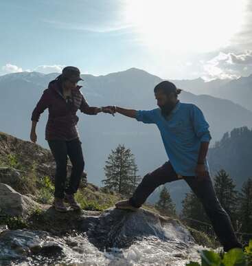 Un homme aide une femme à traverser un ruisseau rocailleux dans les montagnes, avec en arrière-plan la lumière du soleil et des sommets brumeux, symbolisant le travail d'équipe et l'aventure.