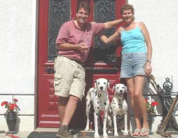 La imagen muestra a un hombre y una mujer delante de una puerta roja, acompañados por dos perros dálmatas. El hombre sostiene un vaso de vino.