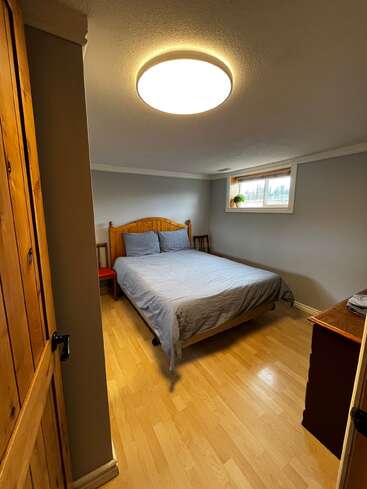 Une petite chambre confortable avec des planchers en bois, un lit en bois, une literie grise, une petite commode, un plafonnier lumineux, une fenêtre avec une plante et de la lumière naturelle.