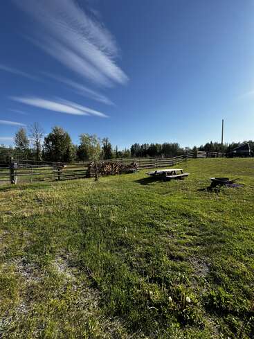 Une vaste cour herbeuse est agrémentée d'une table de pique-nique en bois, d'un foyer, de bois empilé à proximité, de clôtures en bois et d'arbres, le tout sous un ciel bleu clair avec des nuages vaporeux.