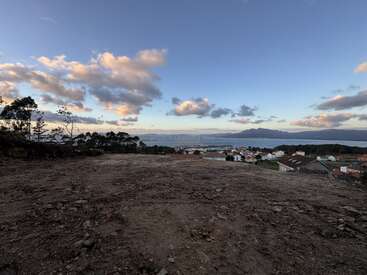 A rocky, barren foreground overlooks a peaceful coastal town under a vast sky with scattered clouds, distant mountains, and calm blue sea in the background.