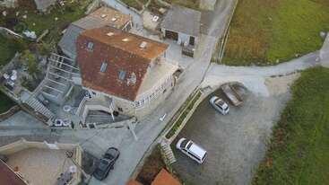 This aerial image shows a rustic house with a red-tiled roof, a small parking lot with two cars, green surroundings, and winding roads nearby.