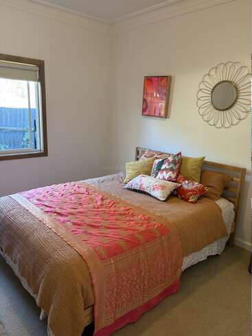 Dieses gemütliche Schlafzimmer hat ein Bett mit bunten Kissen und einer rosa Decke, ein Fenster mit natürlichem Licht und Wandkunst über einem beigen Teppich.