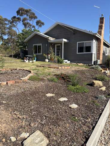Ein graues Haus mit einem gemauerten Schornstein steht unter einem klaren blauen Himmel. Der Hof ist mit Gartenbeeten, Topfpflanzen, Mulch, Trittsteinen und einheimischen Bäumen ausgestattet.