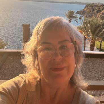 Eine Frau mit kurzen grauen Haaren und Brille macht ein Selfie bei Sonnenuntergang. Hinter ihr liegt eine malerische Aussicht auf das Meer mit Palmen und felsigen Klippen.
