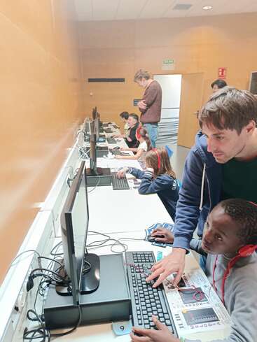 Dans une salle de classe, plusieurs enfants sont assis devant des ordinateurs et travaillent avec attention. Deux adultes se tiennent debout, aidant et supervisant, créant ainsi un environnement d'apprentissage interactif et éducatif pour les enfants.