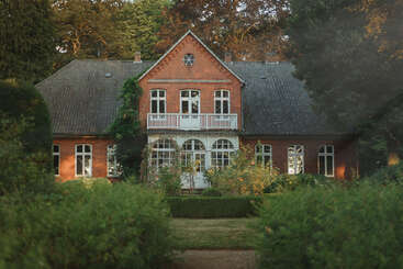 A charming, historic red-brick house with arched windows, a balcony, and lush green gardens, surrounded by tall trees, radiating tranquility and timeless elegance.