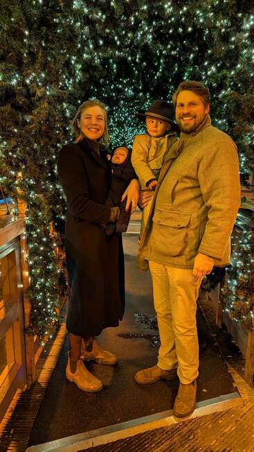 A happy family of four poses under sparkling holiday lights, warmly dressed, smiling and enjoying a festive night together in a beautiful, illuminated tunnel.