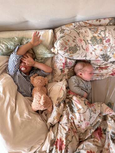 Two young children lie in bed together. The older child has a pacifier, covers their face, and cuddles a teddy bear. Both are cozy in floral bedding.
