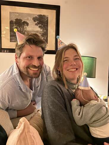 A happy couple, both wearing party hats, smile while sitting closely together. The woman holds a baby, also in a hat, creating a warm, joyful scene.