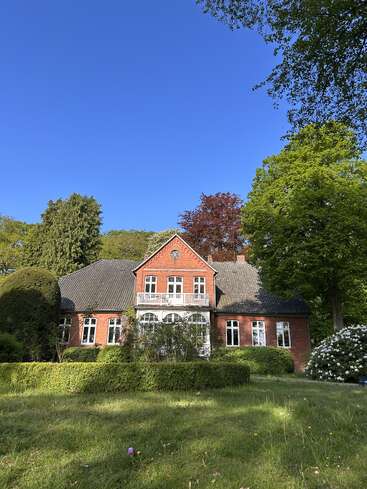 A charming red brick house stands surrounded by lush green bushes, trees, and a well-kept lawn, beneath a vibrant, cloudless blue sky.