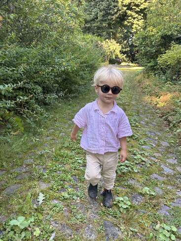 A young child with blonde hair, wearing sunglasses, a pink striped shirt, beige pants, and black boots, walks confidently on a leafy stone path surrounded by greenery.