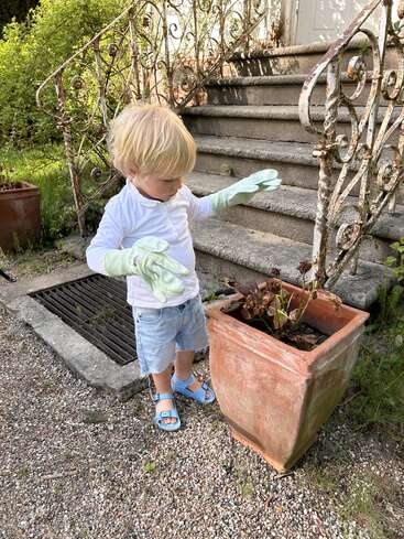 A young child wearing gardening gloves, a white shirt, and blue sandals stands by a large planter near old stone steps, appearing to garden outdoors.