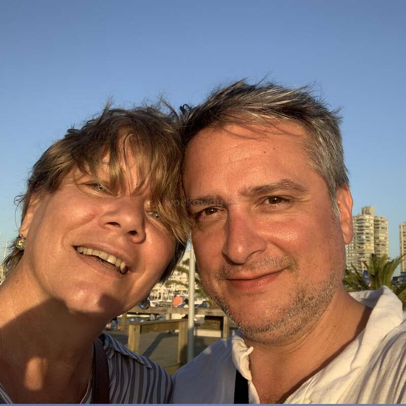 A smiling couple takes a close-up selfie outdoors on a sunny day. Buildings and palm trees are in the background, with clear blue sky above them.