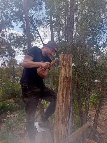 Ein Mann in schwarzem T-Shirt und Mütze hackt draußen mit einer Axt an einem Holzstamm, umgeben von hohen Bäumen und einer natürlichen Buschlandschaft.