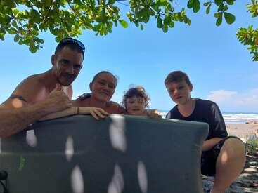 Une famille de quatre personnes sourit sous des branches feuillues sur une plage ensoleillée. Ils sont assis derrière un kayak, profitant de la journée au bord de la mer et du sable.