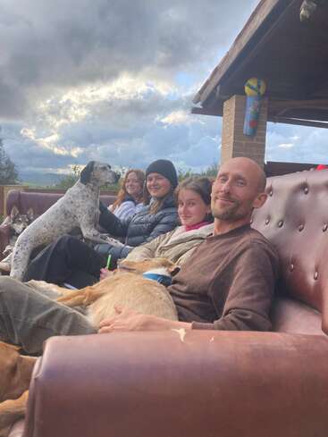 Cuatro personas están sentadas muy juntas en un sofá marrón al aire libre, sonriendo. Varios perros descansan junto a ellas. Un cielo nublado y un ambiente relajante y acogedor completan la escena.