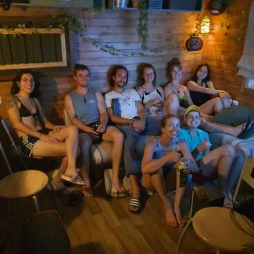 Ocho jóvenes se relajan juntos en una acogedora sala de madera con una cálida iluminación. Sonríen, van vestidos de manera informal y parecen estar disfrutando de la compañía mutua, quizá viendo algo.