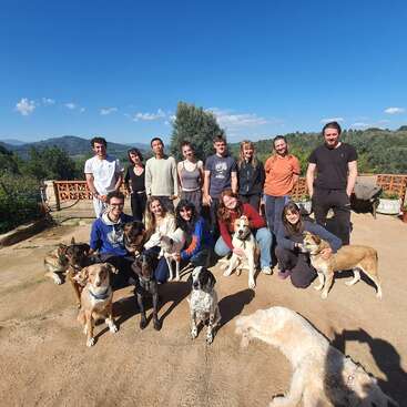 Un grupo de trece personas posa al aire libre en un día soleado, rodeadas por al menos ocho perros. Al fondo se ven montañas, árboles y un cielo azul y despejado.