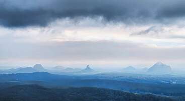 Eine neblige Landschaft zeigt einzigartige, rund geformte Berge, die sich unter dramatischen, dunklen Wolken aus einer bewaldeten Ebene erheben. Sanfte Blautöne dominieren und schaffen einen ruhigen, stimmungsvollen Ausblick.