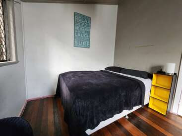 Das Bild zeigt ein schlichtes Schlafzimmer mit einem Bett, das mit einer schwarzen Decke bedeckt ist, einem gelben Nachttisch, Holzböden und einem motivierenden Wandbild über dem Bett.