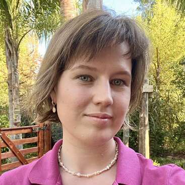 Une femme aux cheveux bruns courts et aux yeux verts se trouve à l'extérieur ; elle porte un chemisier rose, un collier de perles et des boucles d'oreilles. On aperçoit des palmiers et une clôture en bois.