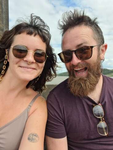 The image depicts a man and woman posing for a selfie, with the woman on the left and the man on the right, both wearing sunglasses and smiling at the camera.