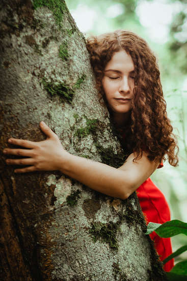 Une femme aux longs cheveux bouclés enlace un tronc d'arbre, le visage serein et les yeux fermés, vêtue d'une robe rouge, dans un cadre naturel avec de la verdure en arrière-plan.