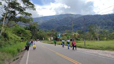 Un groupe de personnes marche le long d'une route de campagne, entouré d'une végétation luxuriante et de montagnes, sous un ciel partiellement nuageux, évoquant un paysage champêtre pittoresque et paisible.
