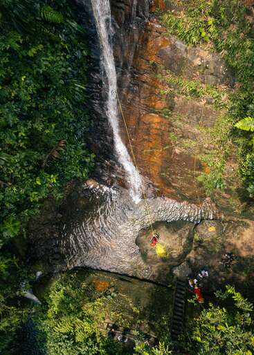 Une personne descend en rappel le long d'une cascade, au milieu d'une forêt luxuriante, avec des cordes, des rochers, l'eau en contrebas et un groupe de personnes qui l'observent à proximité.