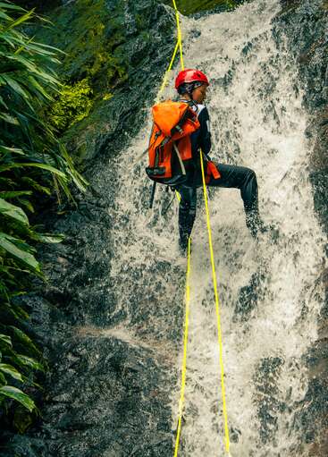 Une personne portant un casque rouge et un sac à dos orange descend en rappel le long d'une cascade rocheuse à l'aide de cordes jaunes, au milieu d'une végétation luxuriante et des éclaboussures d'eau. Scène d'aventure.
