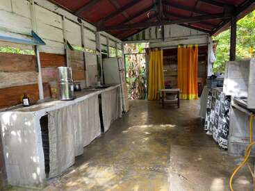 Il s'agit d'une cuisine rustique en plein air avec des comptoirs en béton, des rideaux en tissu, des appareils de base et un toit en métal. Des rideaux jaunes séparent l'espace, entouré de verdure.