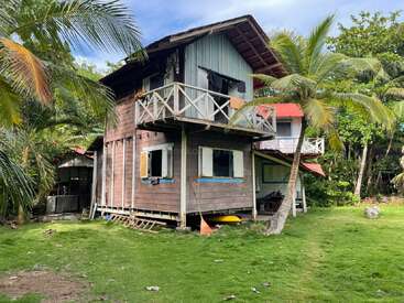 Cette image montre une maison rustique en bois de deux étages avec un balcon, entourée de palmiers et de verdure, sous un ciel lumineux, évoquant une atmosphère tropicale et détendue.