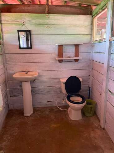 Cette salle de bains rustique se compose de toilettes simples, d'un lavabo sur pied, d'un petit miroir, d'une étagère en bois, d'une poubelle verte, de murs en planches de bois, d'un sol en béton et d'une fenêtre ouverte.