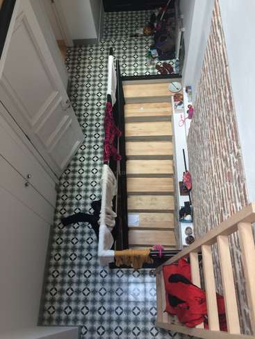 La imagen muestra una escalera con peldaños de madera y barandilla negra, situada en un pasillo de paredes blancas y azulejos estampados, con diversos objetos esparcidos por el suelo y los escalones.
