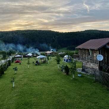 Une scène champêtre paisible avec des tentes, des kiosques et une cabane rustique en bois. Une pelouse d'un vert luxuriant, la fumée des feux de camp et une forêt en toile de fond, le tout sous un ciel spectaculaire.