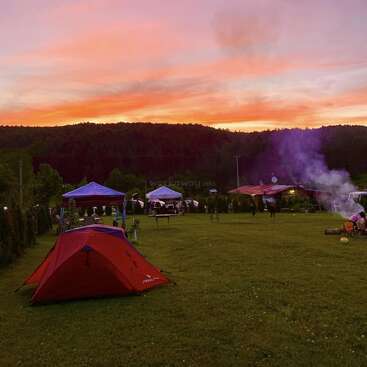 A vibrant sunset sky lights up a grassy campsite. Several tents and canopies are set up. Smoke rises from a campfire while people gather nearby, enjoying nature.