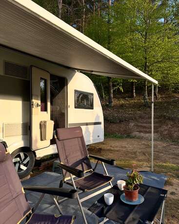 Un coin de camping chaleureux avec une caravane sous un auvent, deux chaises, une petite table avec des tasses à café et une plante en pot, le tout entouré d'arbres.
