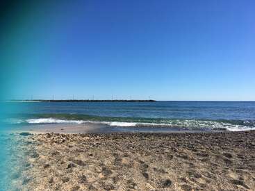 The image depicts a serene beach scene with a sandy shore, gentle waves, and a clear blue sky, featuring a distant pier or jetty in the background.