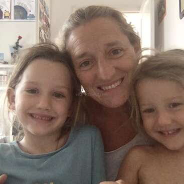 Eine lächelnde Frau posiert eng mit zwei kleinen Kindern in einem Raum. Das Trio sieht glücklich aus und teilt einen warmen Moment in einem gemütlichen, gut beleuchteten Raum.