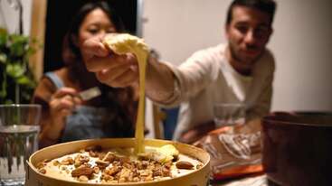 Two people are enjoying a meal together. One person is dipping bread into a gooey, melted cheese topped with nuts. The atmosphere feels cozy and casual.