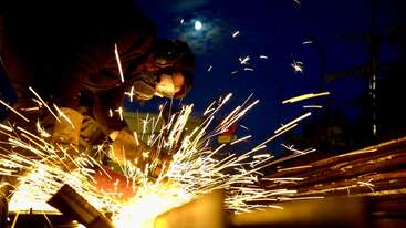 A worker in protective gear uses a grinder at night, creating a burst of bright sparks against the dark blue sky, illuminated by the moon above.