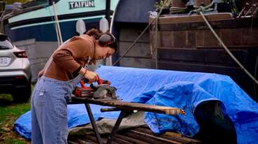A person wearing earmuffs and overalls uses a circular saw to cut wood outdoors. A boat named "TAIFUN" and a blue tarp are visible behind them.