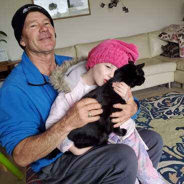 A imagem mostra um homem sentado no chão, segurando uma menina e um gato preto, com um sofá e um tapete ao fundo.