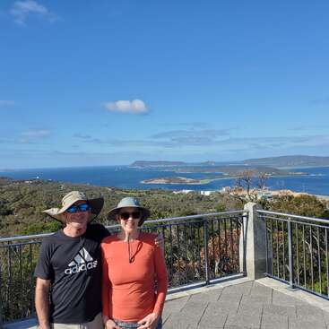 A imagem mostra um homem e uma mulher em pé em uma varanda com vista para um corpo de água sereno, cercado por uma vegetação exuberante e um céu azul claro.