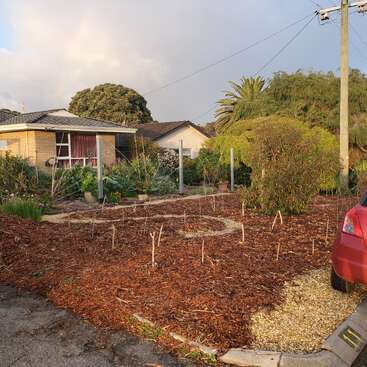A imagem mostra uma pequena casa de um andar com um jardim e um carro vermelho estacionado na frente, cercada por árvores e um céu nublado.