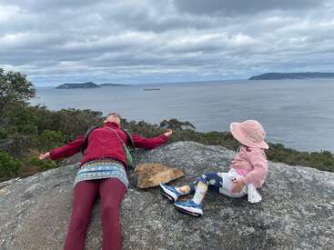 A imagem retrata uma cena serena de uma mulher e uma criança sentadas em uma rocha com vista para um corpo d'água, com um céu nublado acima.