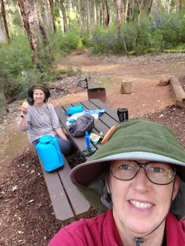 A imagem mostra duas mulheres em uma área arborizada, uma sentada em uma mesa de piquenique com equipamento de acampamento e a outra em primeiro plano usando um chapéu e uma camisa vermelha.