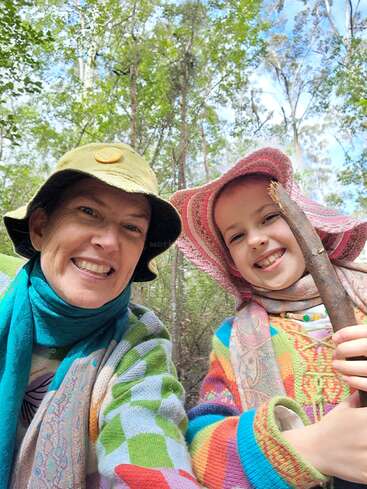 Une femme souriante et un enfant portant des pulls et des chapeaux colorés prennent un selfie dans une forêt verdoyante. L'enfant tient un bâton, les deux ont l'air joyeux.